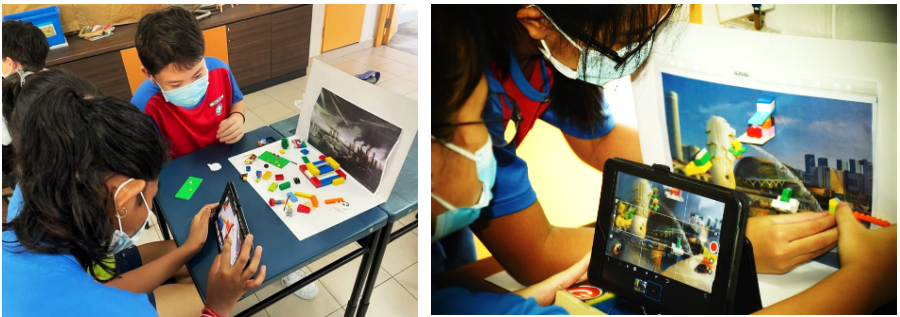 Two masked children use a tablet to view colorful LEGO blocks arranged on a table next to a printed photo.